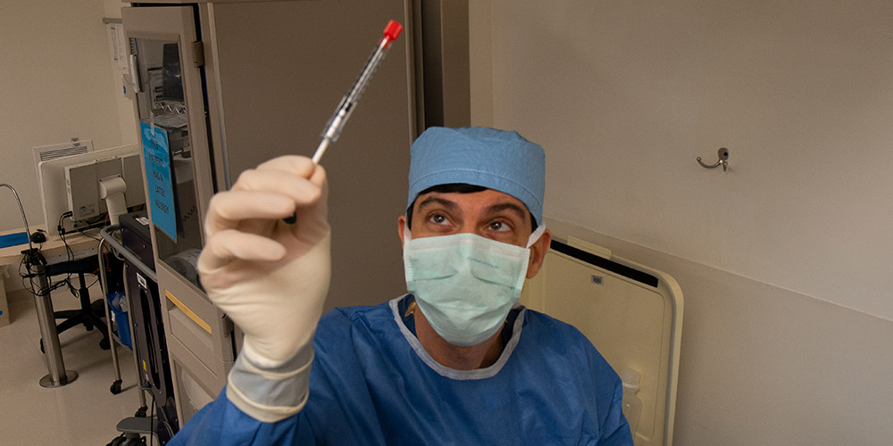A doctor in scrubs, surgical mask, and gloves holds up a test tube
