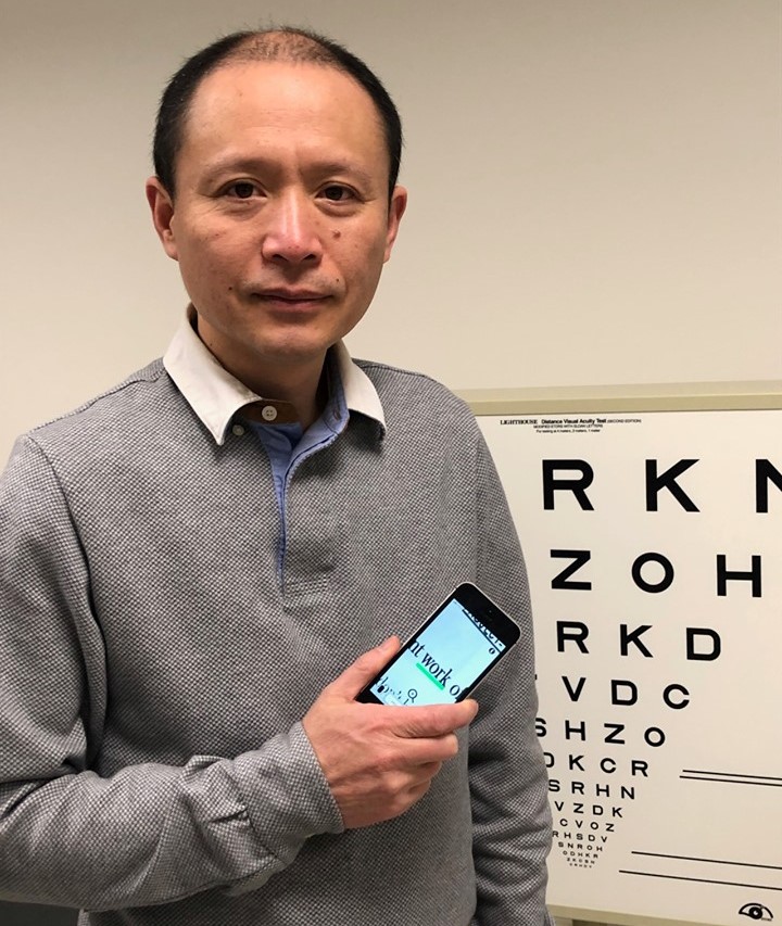 Dr. Gang Luo in front of eye poster holding smart phone with vision app